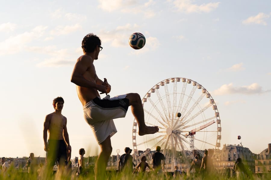 Un joueur reprend le ballon de l'extérieur du pied dans un parc parisien au coucher du soleil, la grande roue en arrière-plan.